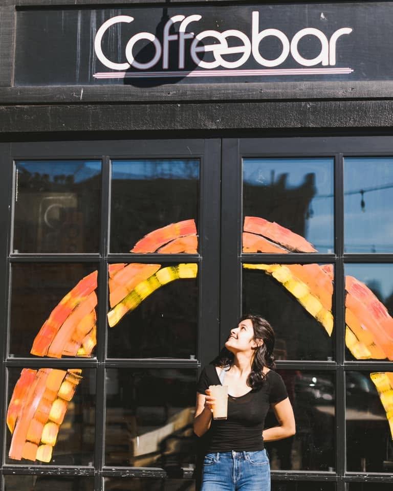 Woman standing outside a coffee shop with a rainbow painted on the window.