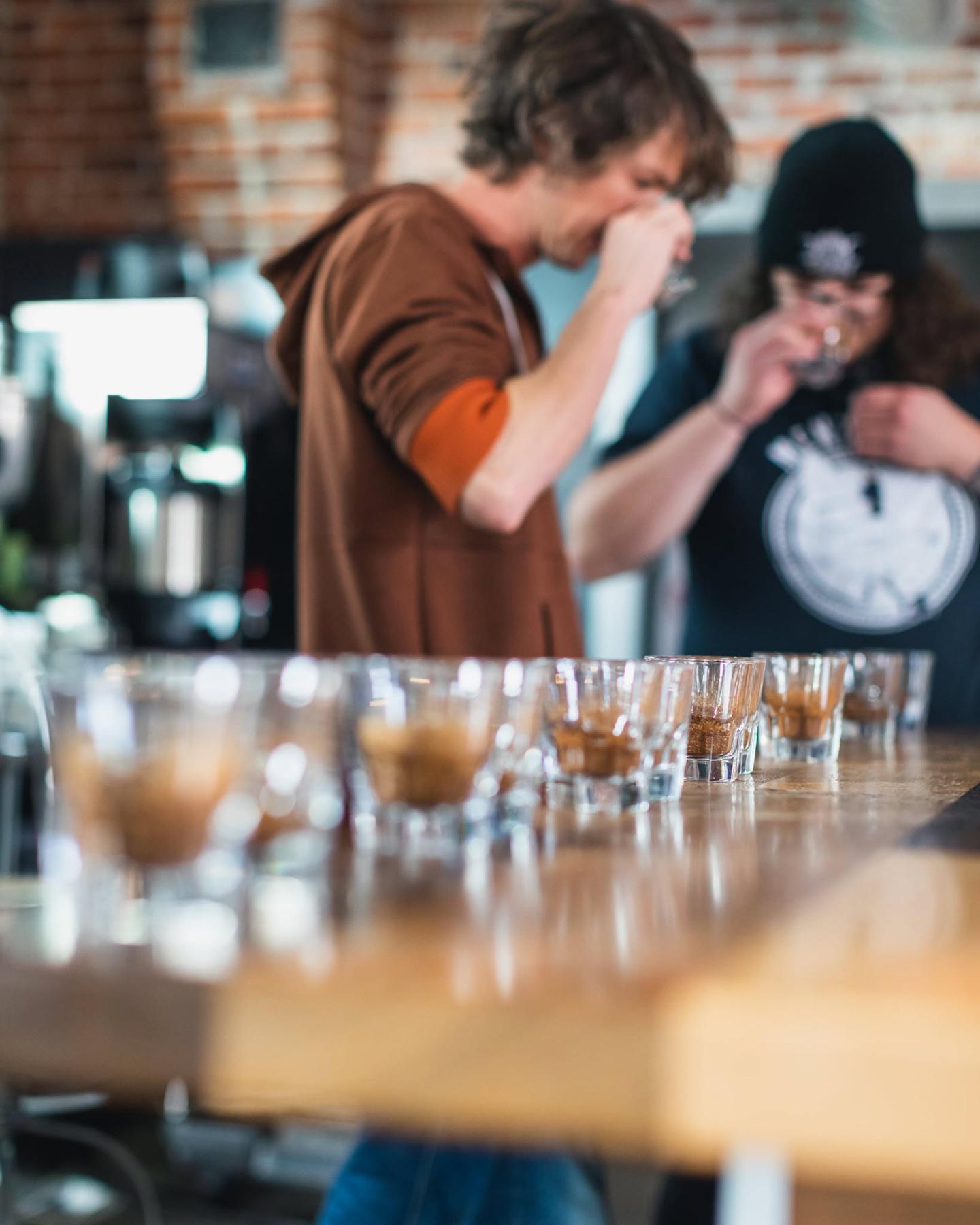 Two people tasting coffee from glasses on a wooden table.