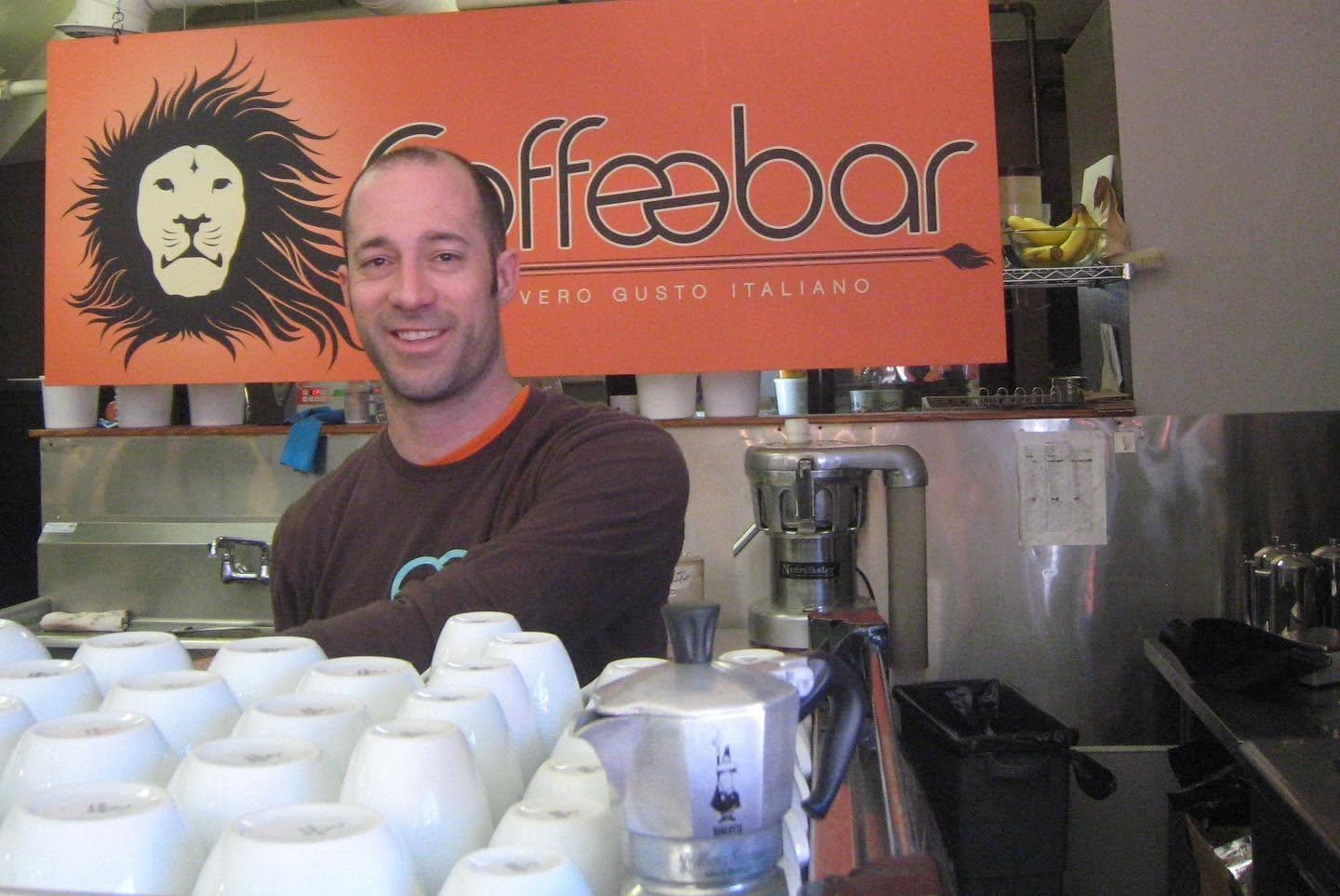 Barista smiling behind coffee cups and espresso machine, with orange coffeebar sign.