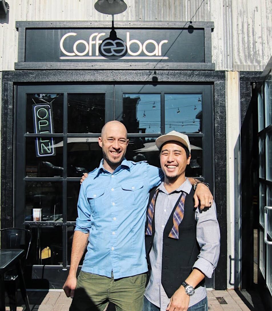 Two men smiling in front of a coffee shop entrance.