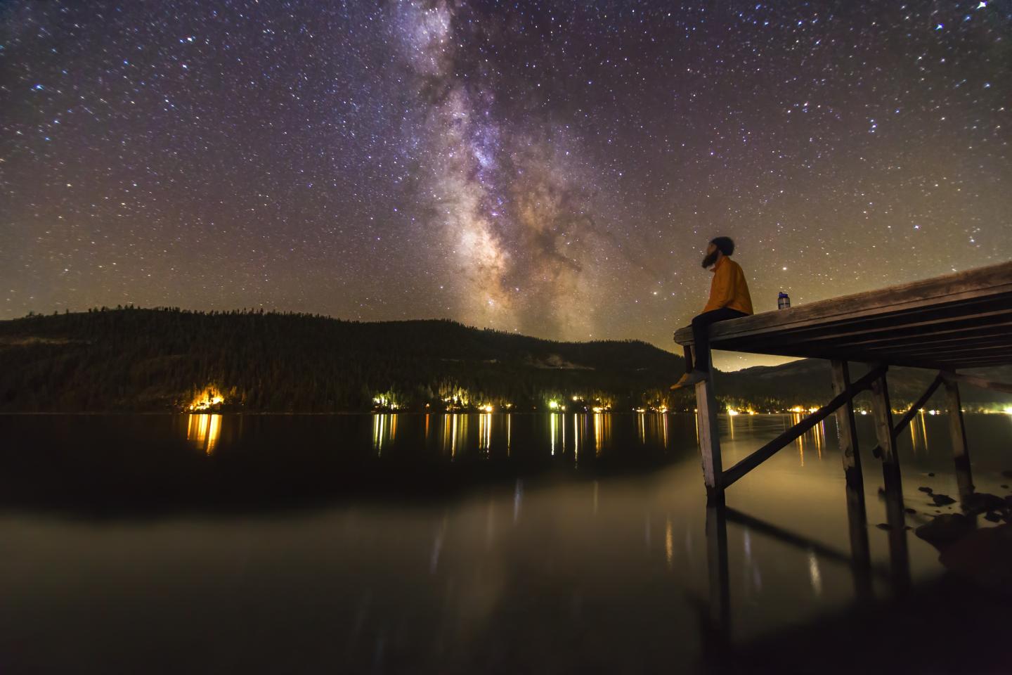 Person on pier gazing at starry sky with Milky Way over calm lake.