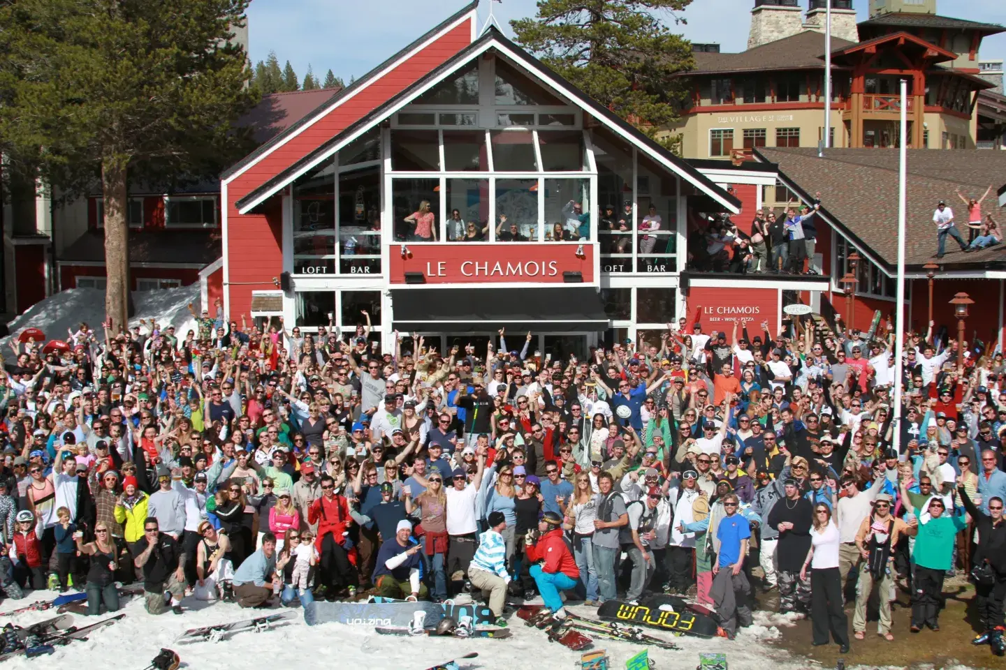 Large crowd gathered in front of a red chalet, some standing on snow.