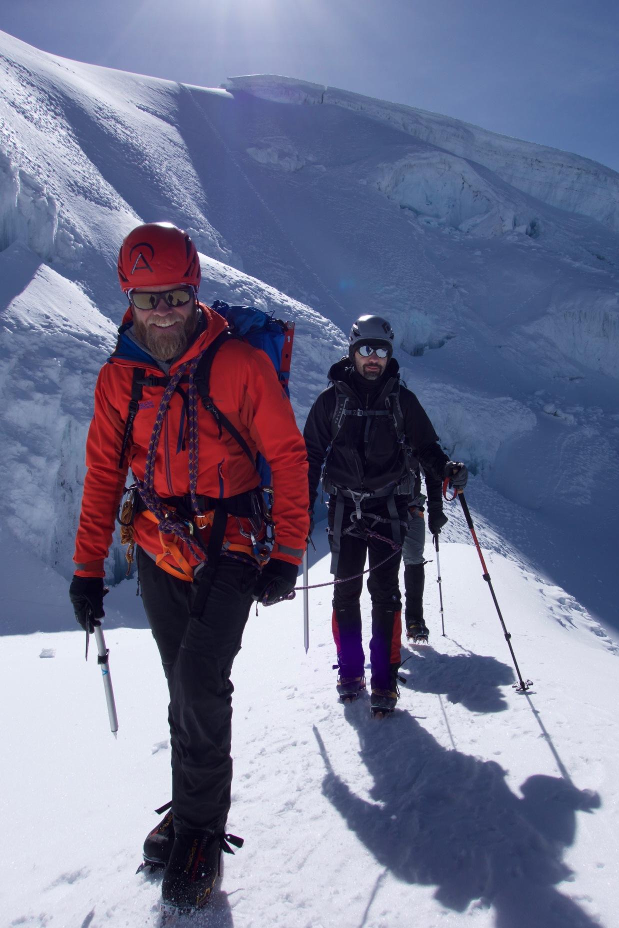 Two climbers in snow gear ascend a sunny, snowy mountain ridge.