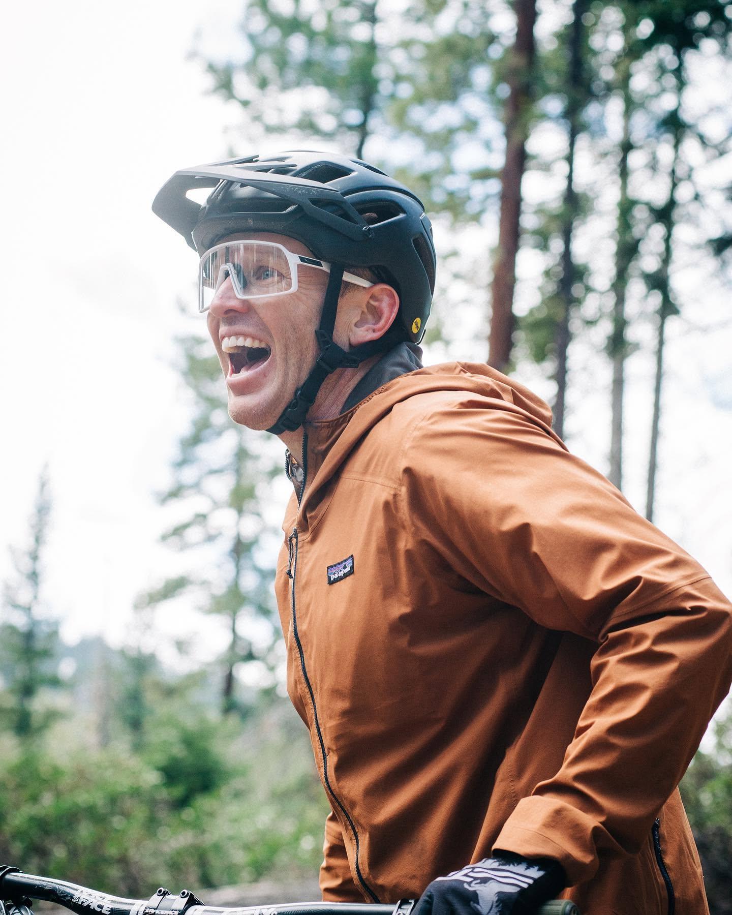 Man in brown jacket and helmet, smiling in a forest setting.