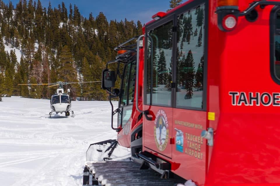 Red snowcat and helicopter on snowy mountain terrain with trees in the background.