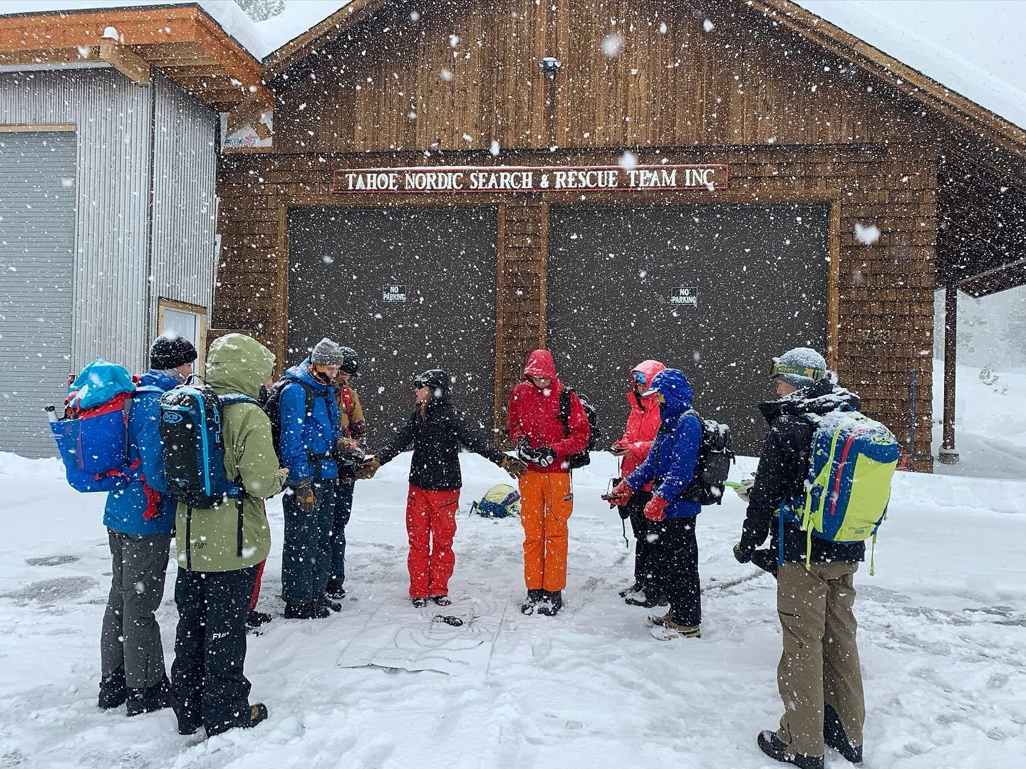Group of people in winter gear standing in a snowy circle near a wooden building.