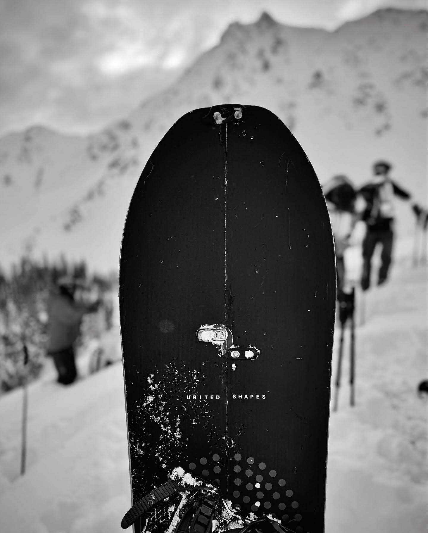Snowboard in foreground with skiers in snowy mountain background, black and white.