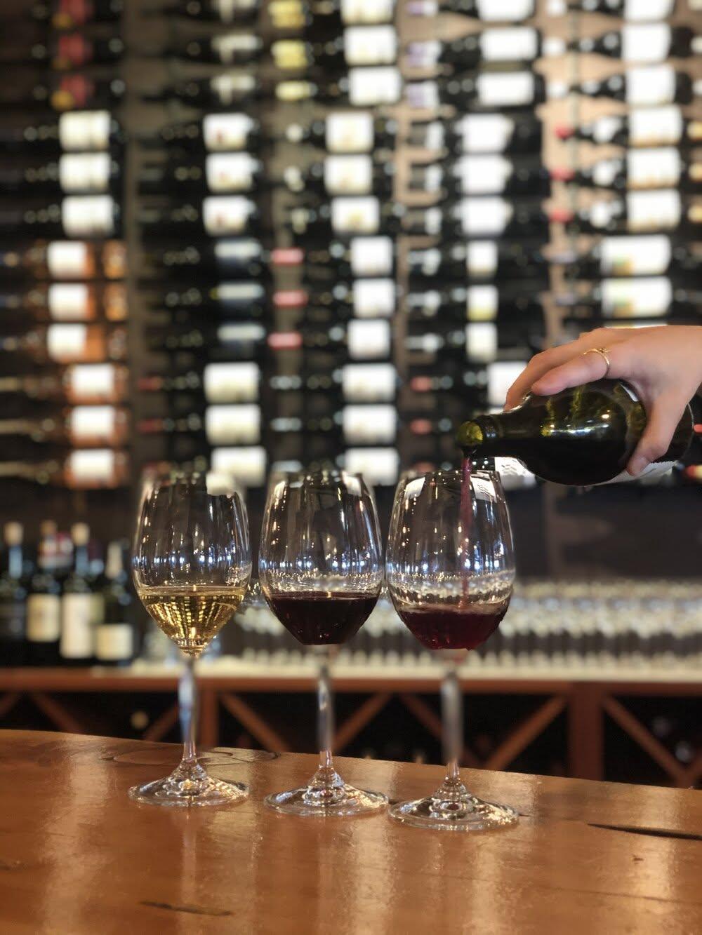 Three wine glasses being filled on a bar, wine bottles displayed in the background.
