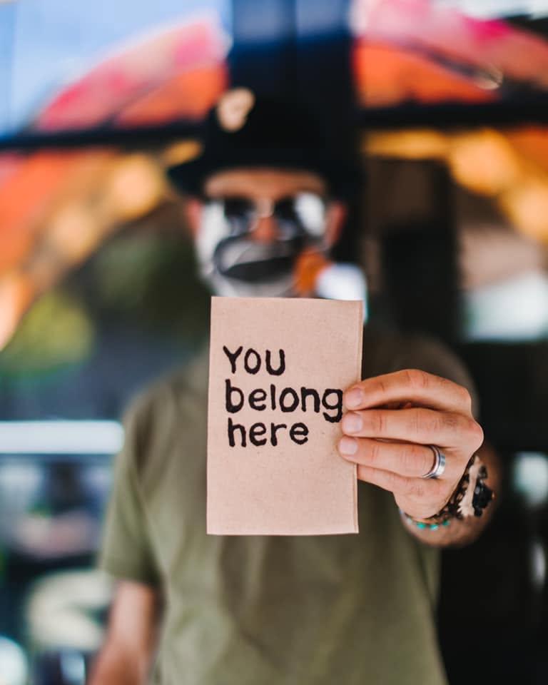 Man holding a card with "You belong here" text. Blurred colorful background.