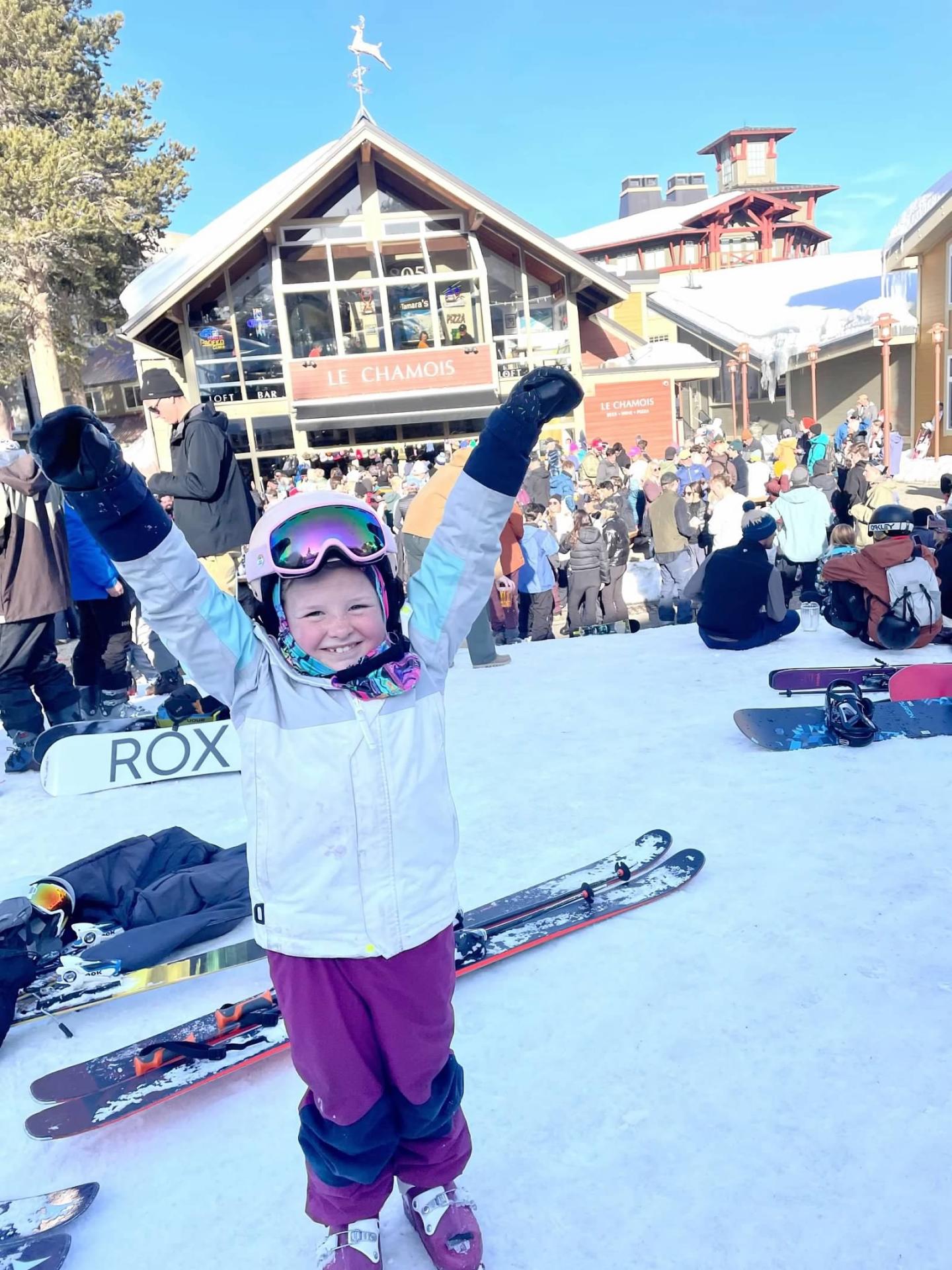 Child in ski gear, smiling with arms raised, snowy mountain lodge background.