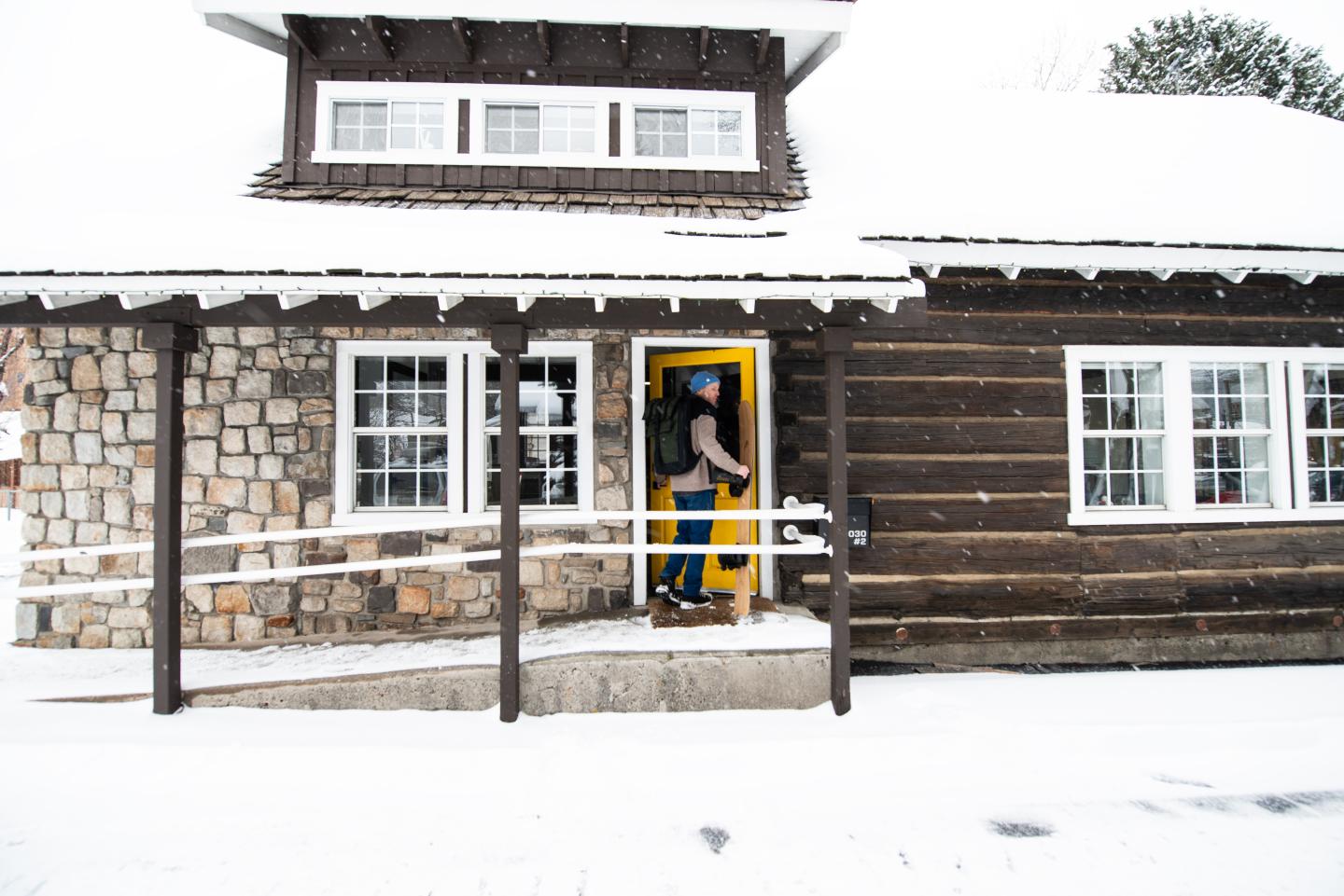 Snow-covered cabin with a person entering through a yellow door.