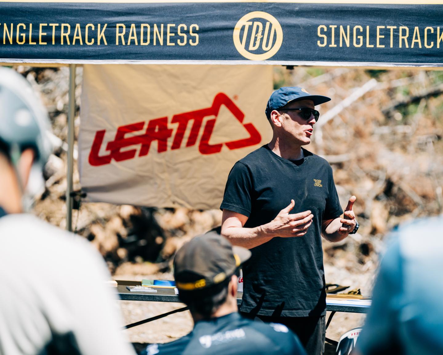 Man speaking outdoors at an event with people listening attentively.