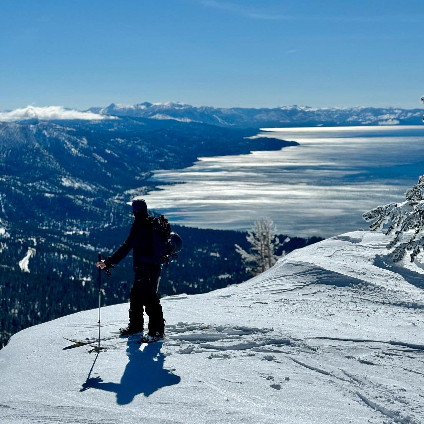 Skier on snowy mountain overlooking a lake under clear blue sky.