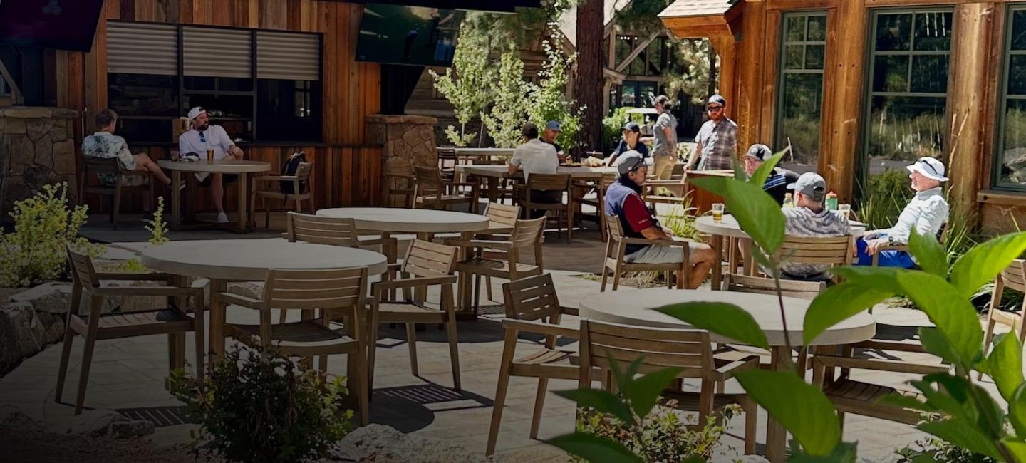 Outdoor patio with wooden tables, chairs, and people dining under sunlight.