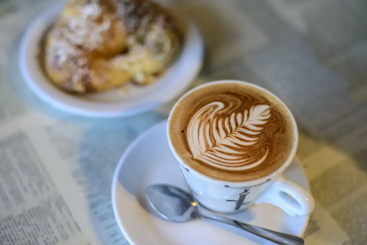 Latte with fern latte art, alongside a croissant on a newspaper-covered table.