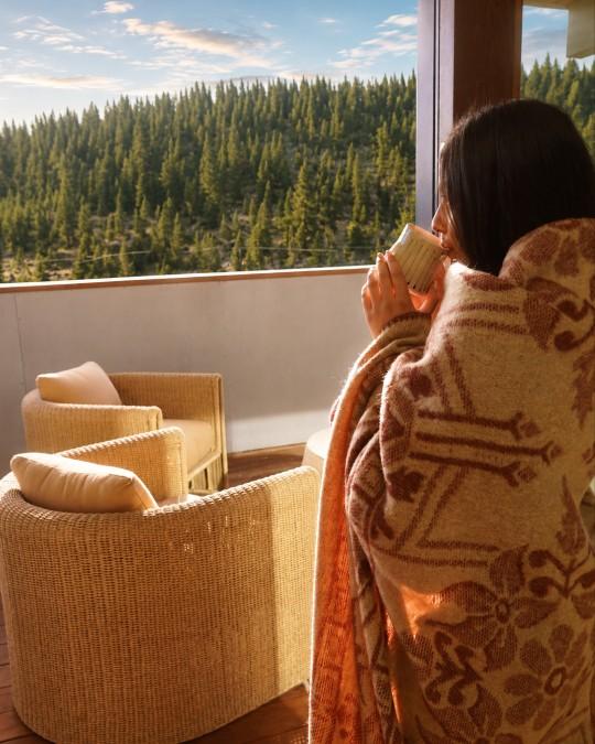 Woman wrapped in a blanket sipping a drink on a balcony with forest view.
