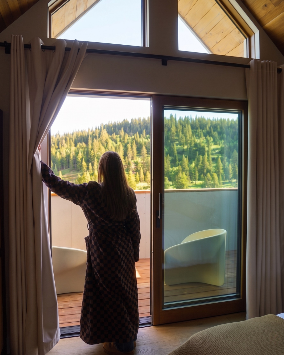 Woman in robe opens curtains, views forested landscape through glass door.