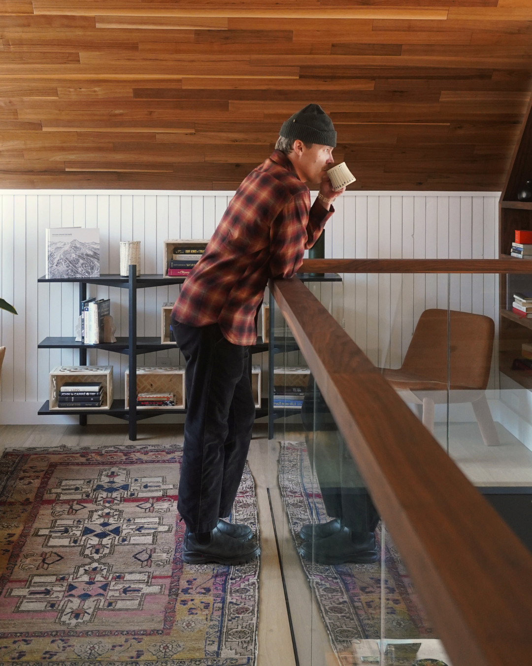 Person in beanie and plaid shirt, sipping from mug, leaning on railing in cozy, wooden room.