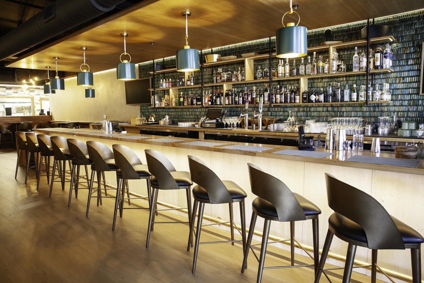 Modern bar interior with a long counter, stools, pendant lights, and bottle shelves.