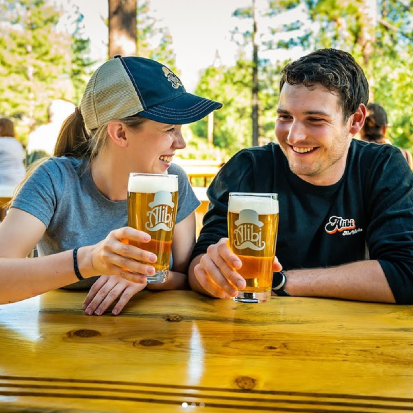 Two friends smiling and clinking beer glasses at a sunny outdoor table.