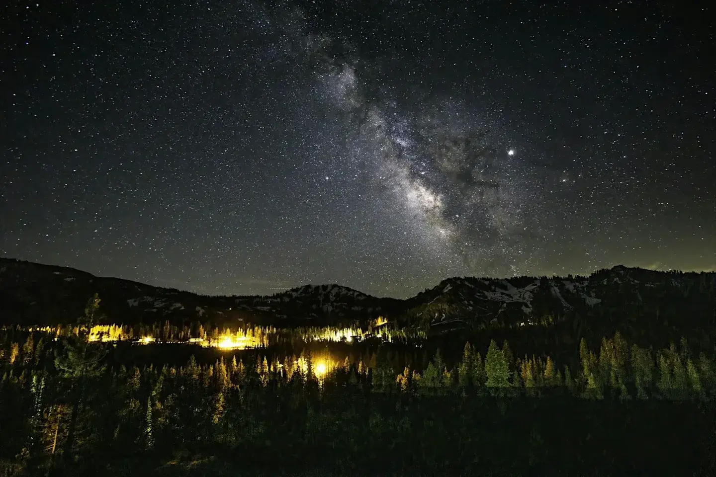 Milky Way over a dark forest and mountain ridge, with warm lights below.