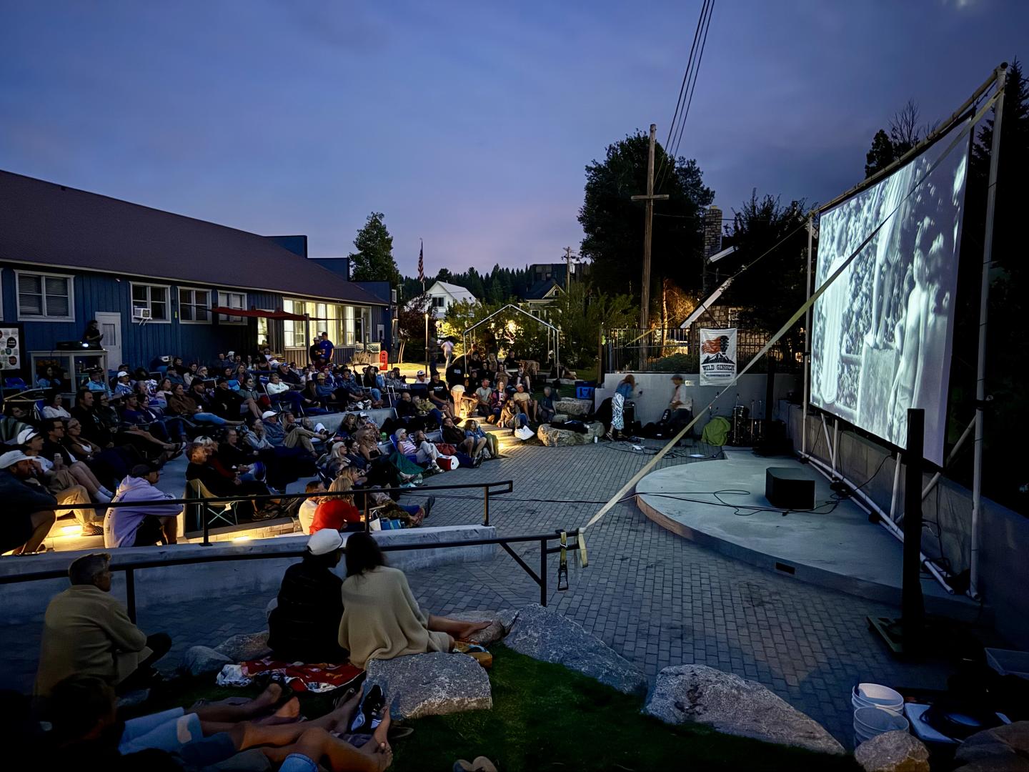 Outdoor movie screening at dusk with people seated on blankets facing a large screen.
