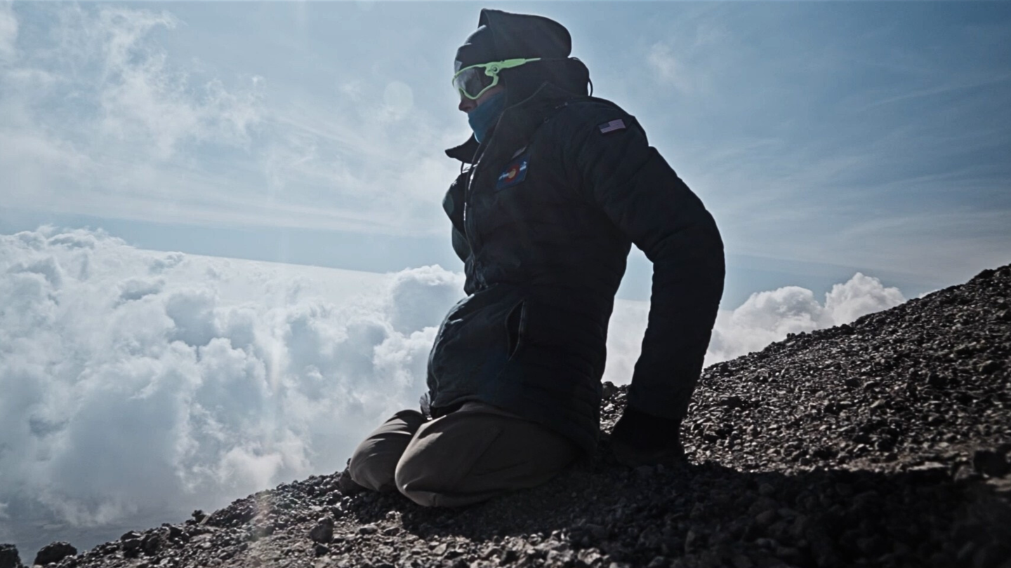 Hiker kneeling on a rocky mountain ridge above clouds at sunrise.