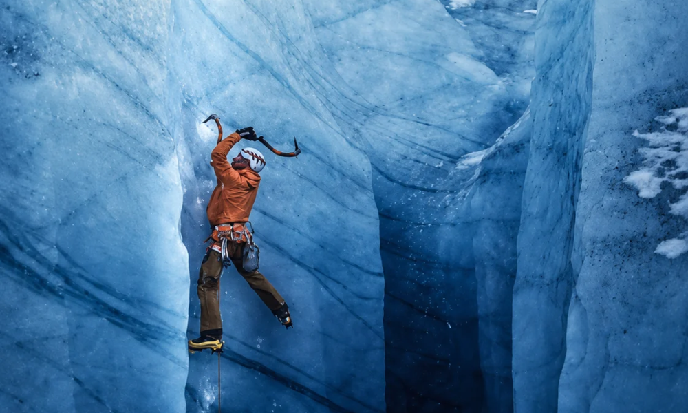 Climber in orange jacket ascending a narrow blue ice crevasse.