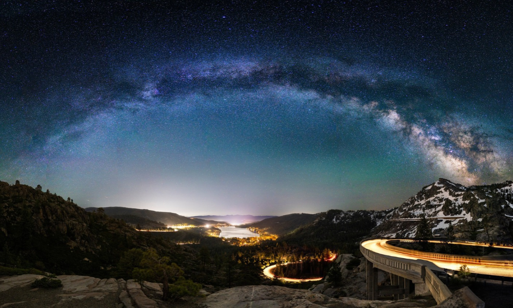 Milky Way arches over a lit mountain road and valley at night.