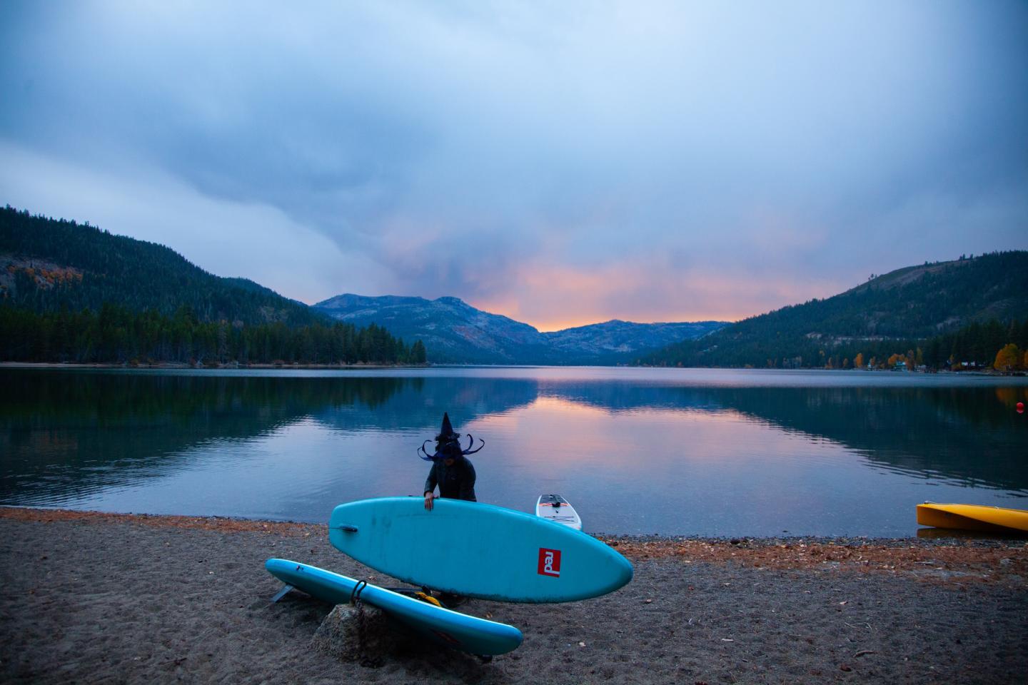 Person standing beside a turquoise paddleboard on a calm mountain lake at sunset.