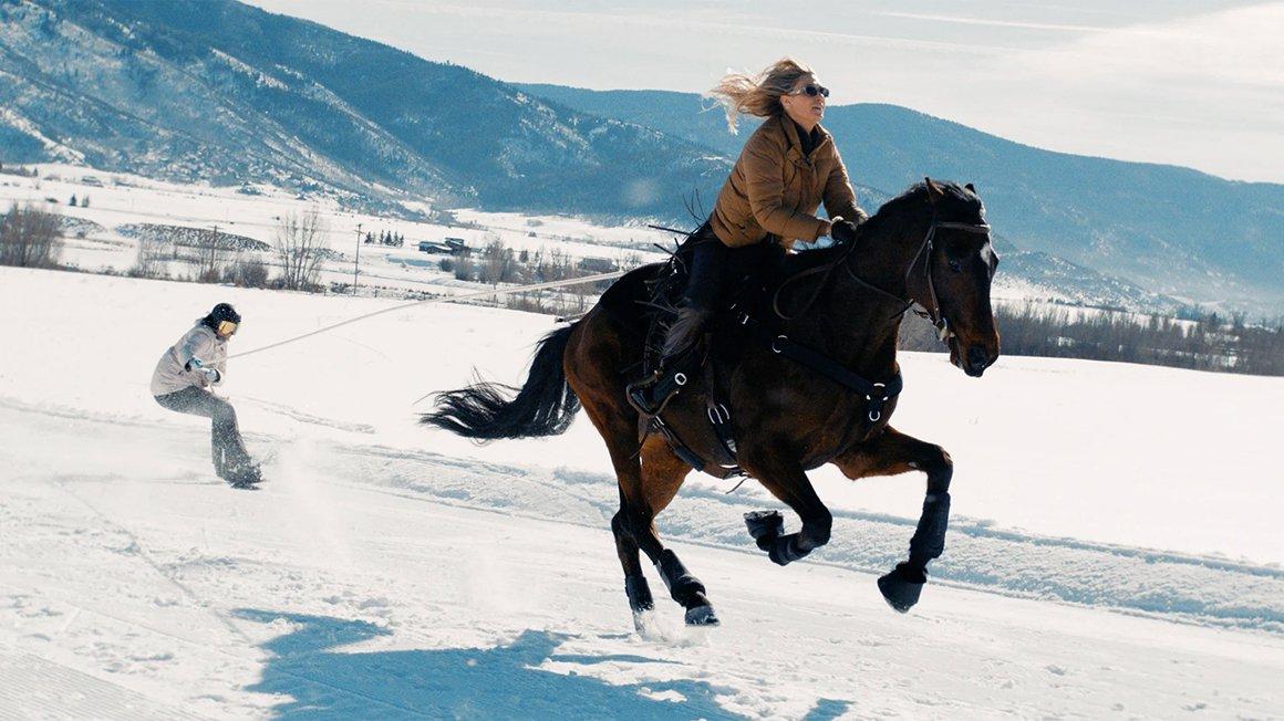 Rider on a rearing black horse in a snowy field, with a skier behind.