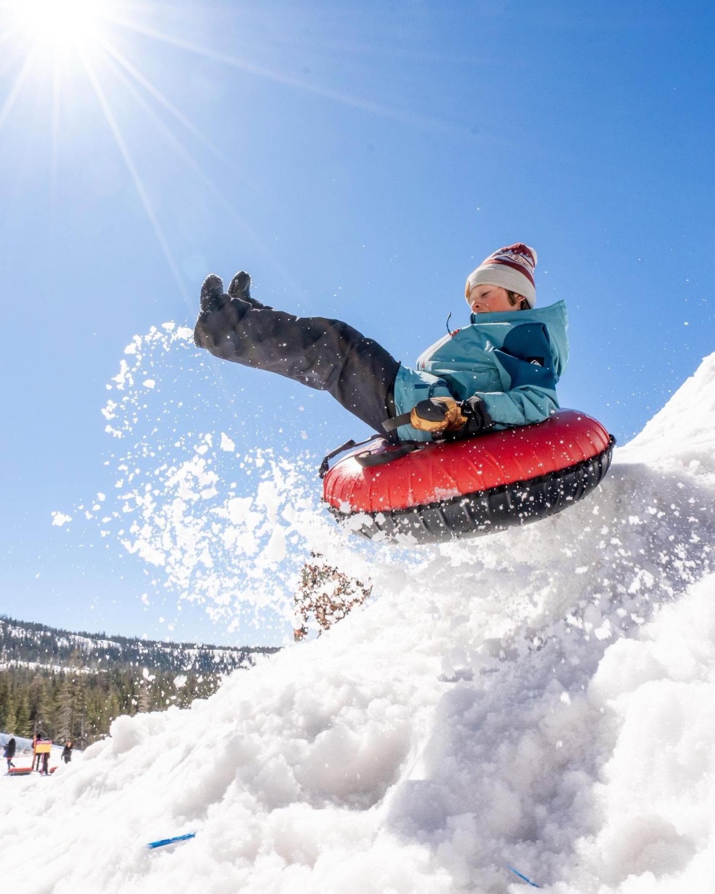 Child in a blue jacket sledding through snow under a bright blue sky.