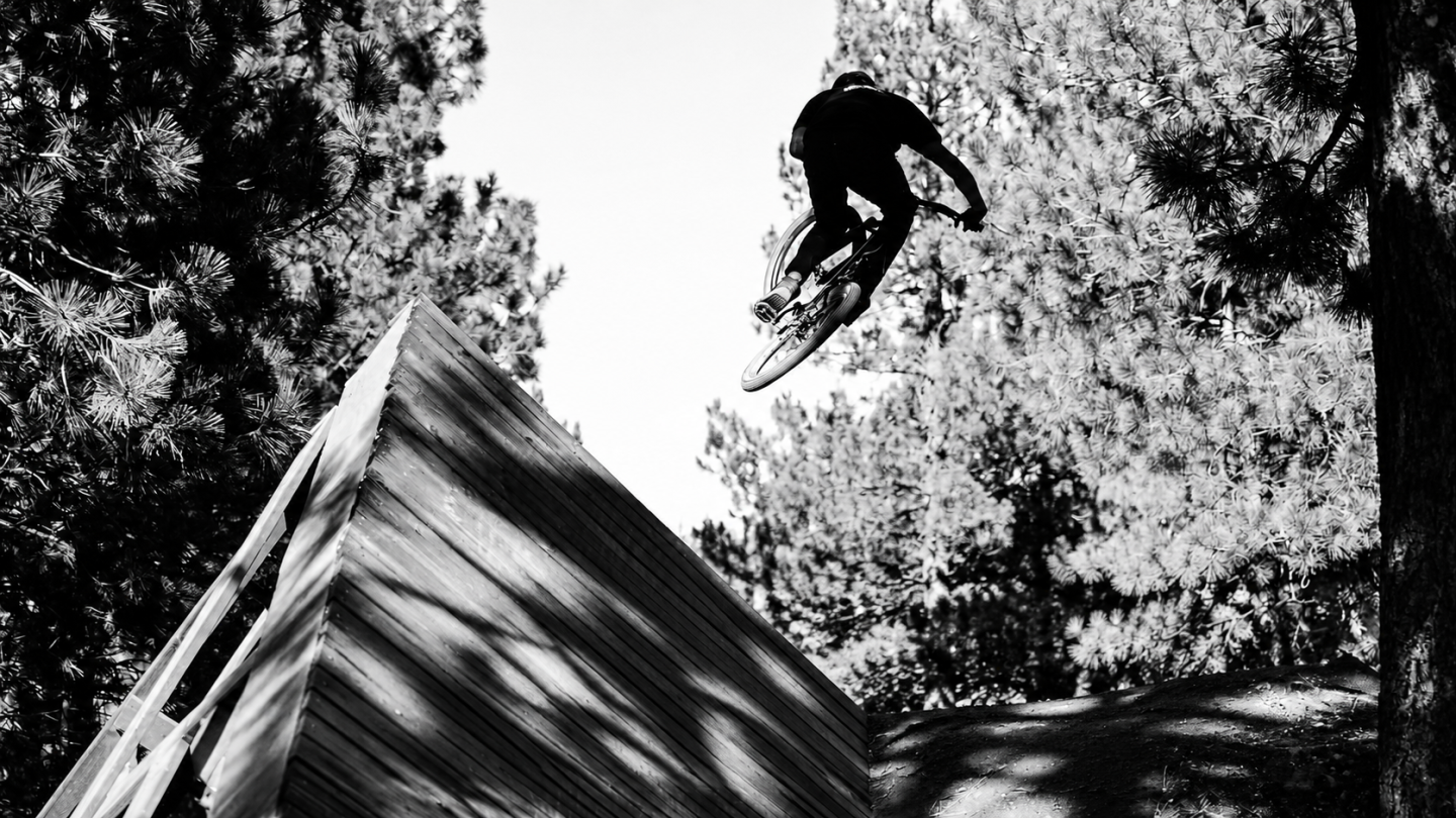 Black-and-white photo of a BMX rider jumping above a wooded trail.