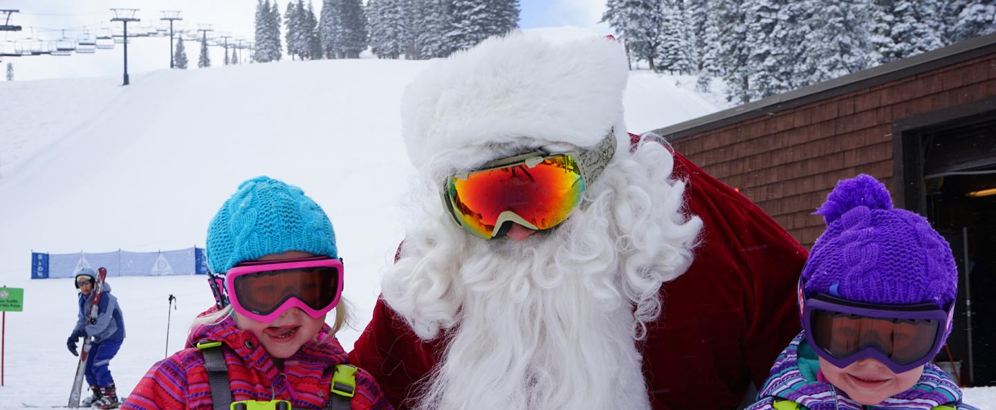 Santa in sunglasses with two children in colorful winter clothes in the snow.