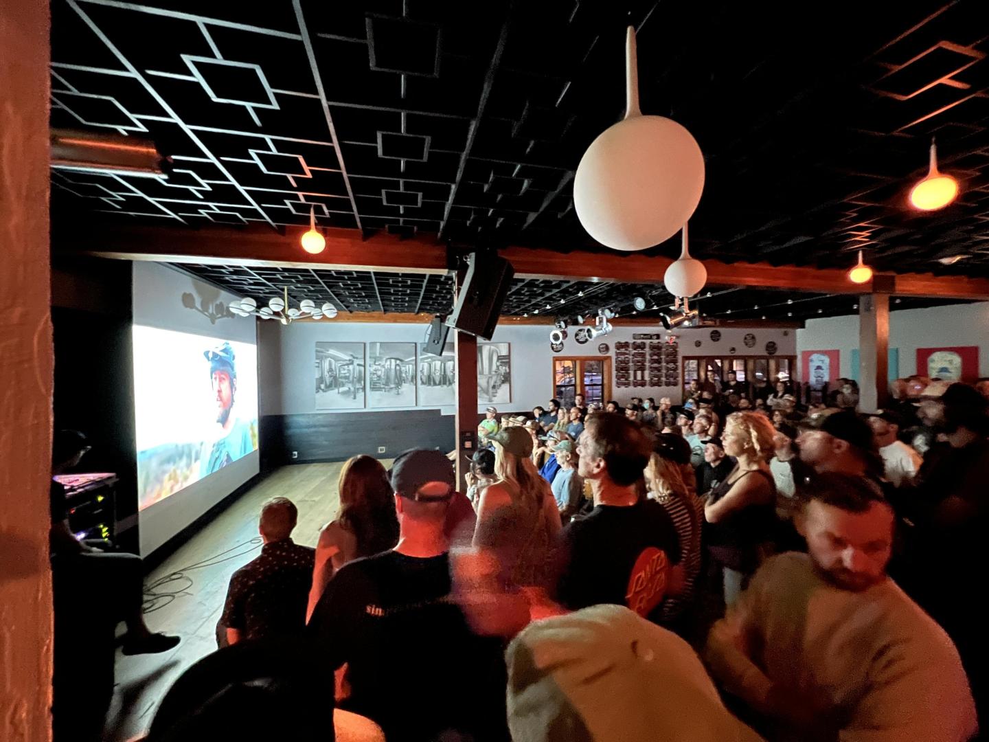 Crowded industrial-style bar with patrons watching a projected screen.