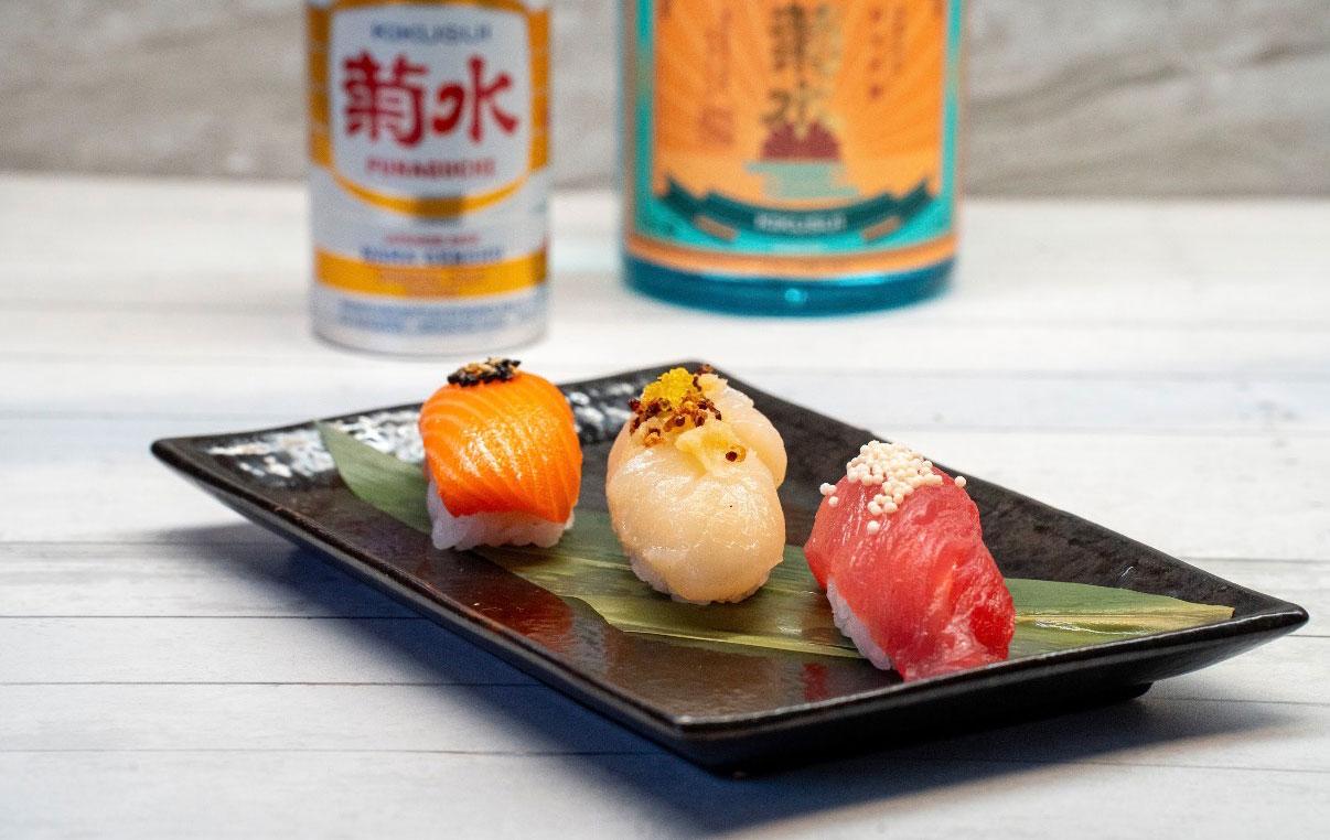 Three pieces of sushi on a black plate, with bottled drinks behind.