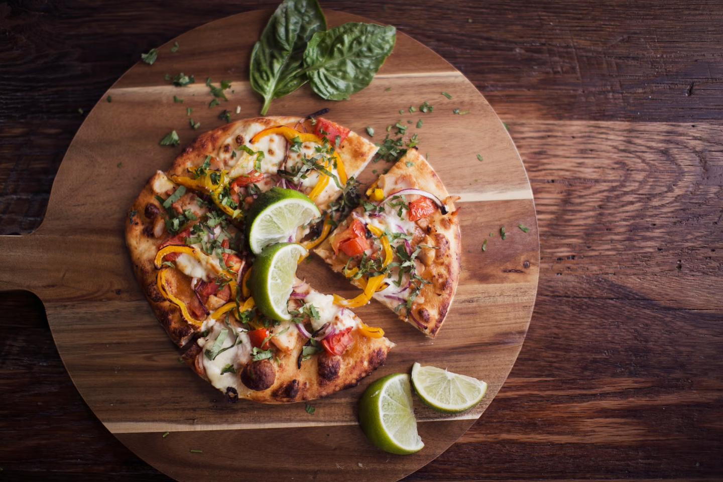 Top-down photograph of a vegetable pizza with lime wedges on a wooden board.